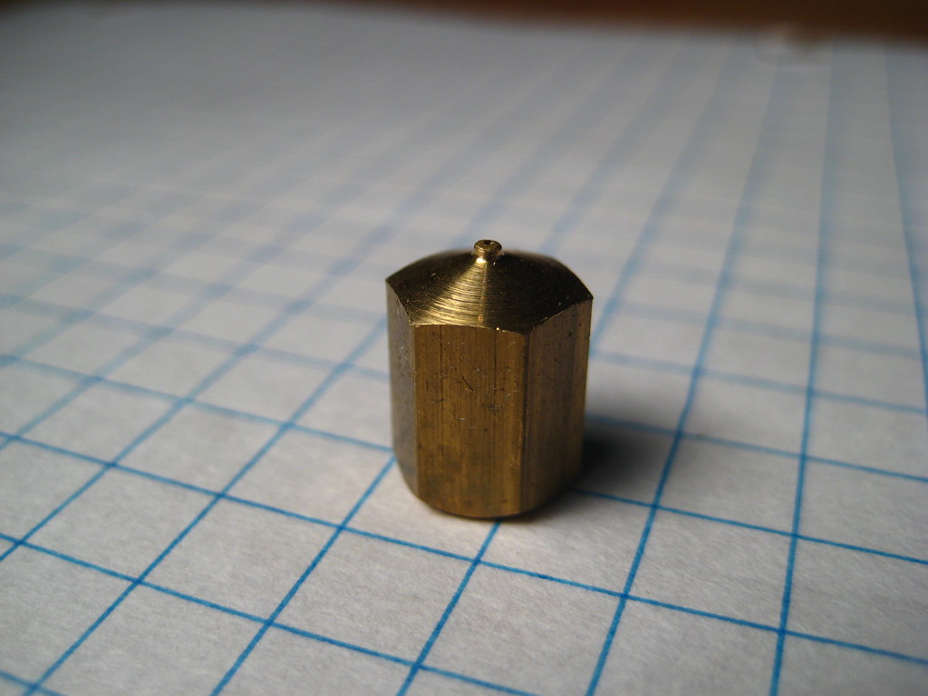 A brass 3D printer nozzle standing on graph paper, the orifice visible at its tip