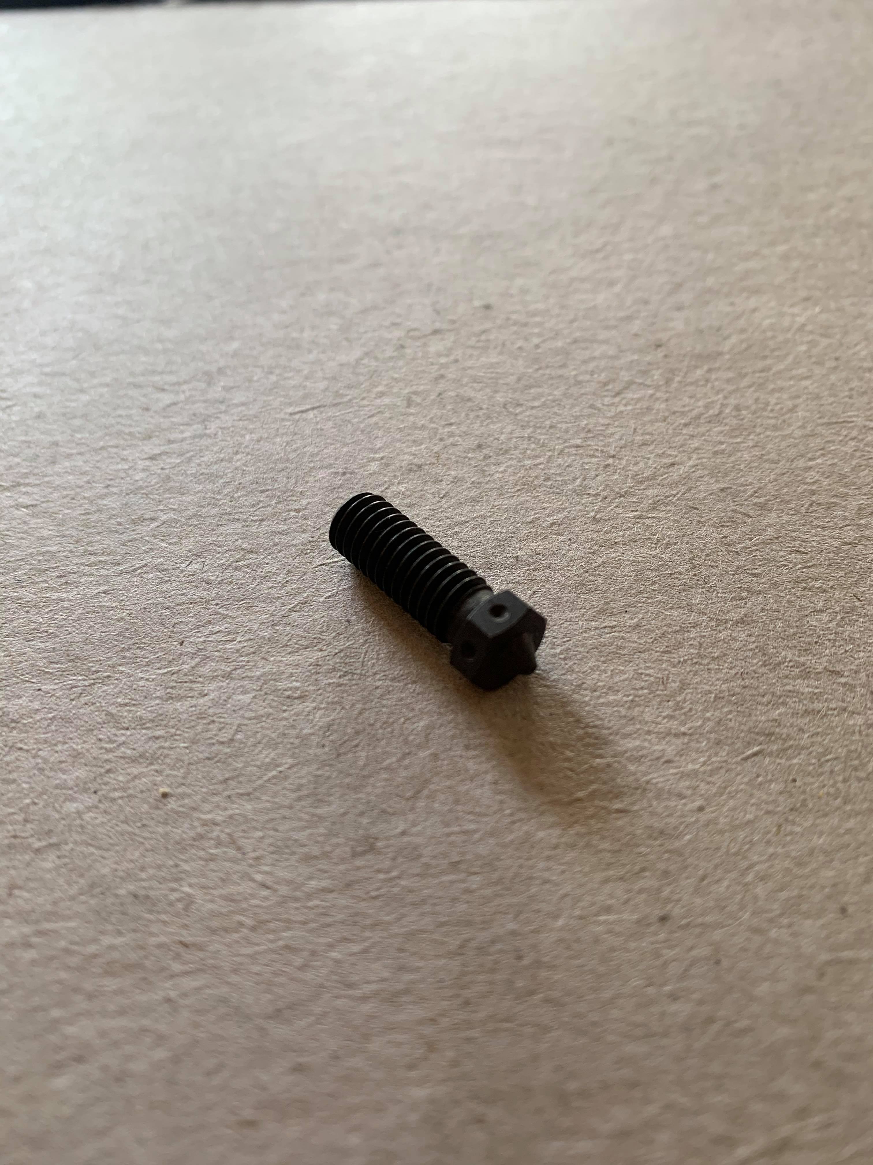 A dark hardened steel 3D printer nozzle lying on a table