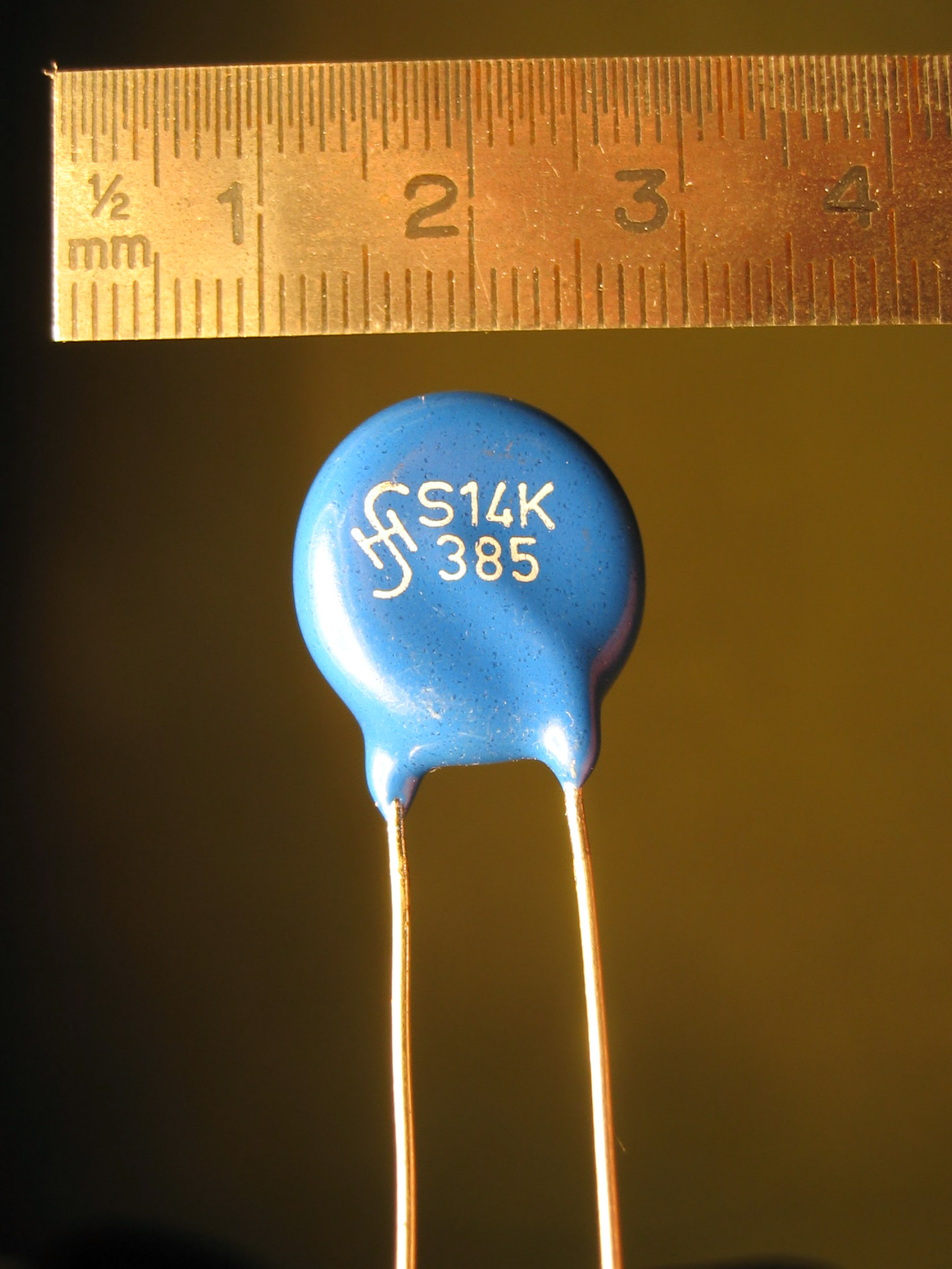 A blue disc varistor with two wire leads, next to a millimetre rule