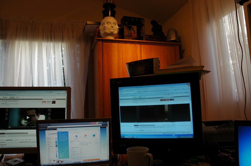 Several monitors on a desk: one shows a video player window with a dark frame, the others show bright browser pages.