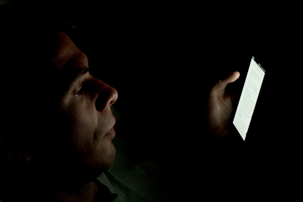 A person in darkness holding a smartphone; only a small patch of the screen with text is lit, the rest of the frame is black.